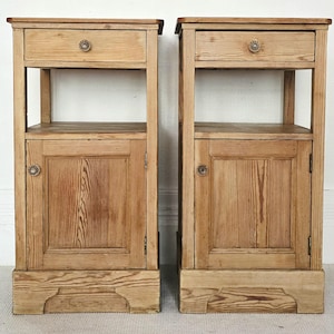 May include: A pair of wooden bedside cabinets. Each cabinet features a drawer, a cupboard door, and an open shelf. The light-coloured wood shows the grain. Decorative knobs adorn the drawer and cupboard door.