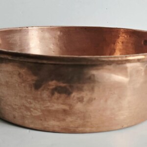 May include: A copper cooking pot with two black handles. The pot has a wide, shallow design and a shiny, reflective surface. The handles are curved and attached to the sides of the pot. This vintage cookware is ideal for cooking and serving.