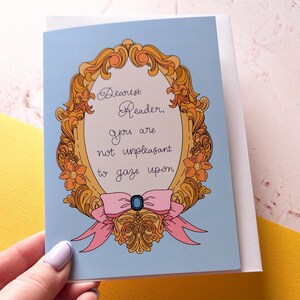 May include: A blue card with a gold ornate frame and a pink bow. The text inside the frame reads "Dearest Reader, you are not unpleasant to gaze upon."