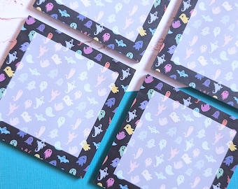 May include: Four square coasters with a black border featuring colorful ghost illustrations. The center of each coaster has a light blue background with more ghost designs. The coasters are arranged on a blue surface.