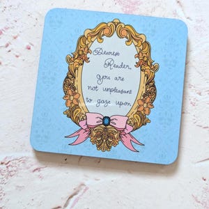 May include: A square coaster with a light blue background and a gold ornate frame. The frame contains the text "Dearest Reader, you are not unpleasant to gaze upon." A pink bow with a blue gem is at the bottom.