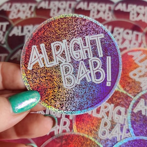 Peut inclure: Un autocollant holographique avec le texte "Alright Bab!" en lettres blanches sur un fond de paillettes arc-en-ciel.