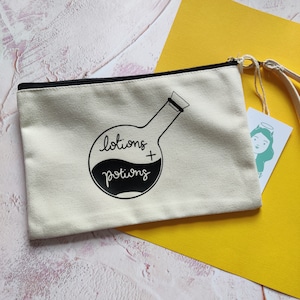 Puede incluir: Una bolsa de lona blanca con cremallera y cordón de ajuste negro. La bolsa tiene una ilustración en blanco y negro de un matraz redondo con el texto "lotions + potions" en ella.
