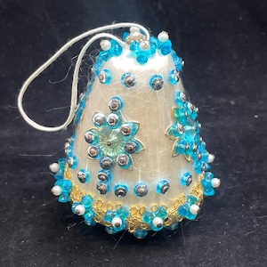 May include: A white bell-shaped Christmas ornament decorated with blue and silver sequins, white beads, and gold trim.