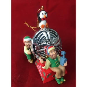 Peut inclure: Ornement de Noël avec un microphone argenté surmonté d'un pingouin avec un nœud papillon rouge. Deux figurines d'elfes encadrent le microphone. L'ornement est posé sur un fond rouge.