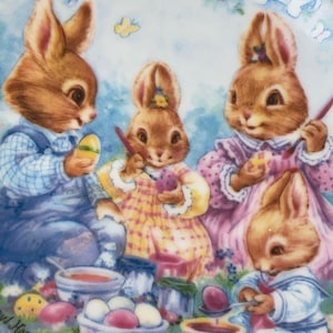 Puede incluir: Un plato vintage de Pascua con cuatro conejos marrones pintando huevos de Pascua. El plato tiene un fondo azul con una pancarta blanca que dice "Easter 1992".