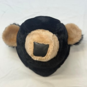 May include: A plush bear head with black fur and tan inner ears and muzzle. The bear's nose is a square of black material. The bear's face is centered in the frame, with a white background.