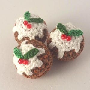 Puede incluir: Tres pudines de Navidad de ganchillo. Cada pudín es marrón con glaseado blanco y decorado con hojas verdes y bayas rojas. Las decoraciones hechas a mano están dispuestas sobre una superficie blanca.