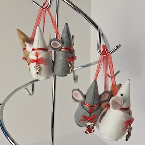 May include: Five felt mouse ornaments hanging on a silver tree stand. The mice are wearing red and white outfits with a candy cane and stocking charm. The ornaments are red and white, grey and white, and brown and white.