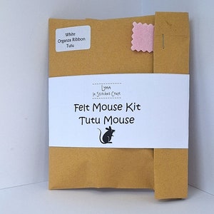 Può includere: Una busta di carta marrone con un'etichetta bianca che dice "Felt Mouse Kit Tutu Mouse" e una piccola illustrazione di topo nero. La busta ha un quadrato di feltro rosa attaccato all'angolo in alto a destra. L'etichetta dice anche "Lynn In Stitches Craft".