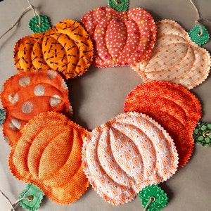 May include: A garland of eight orange and white fabric pumpkins with green stems. The pumpkins are sewn with different patterns and are connected by a twine string.