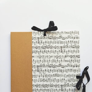 Op de afbeelding: Een bruin notitieboekje met een zwart lint eromheen gebonden. De omslag van het notitieboekje is versierd met een blad muziek.
