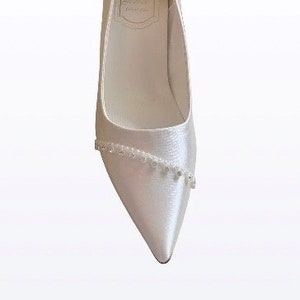 May include: White satin bridal shoes with a pointed toe and a beaded embellishment on the front. The shoes have a small label on the inside with the letter "A" and the text "Atelier" in gold lettering.