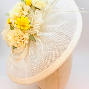 Op de afbeelding: Een witte mesh hoed met een gele en witte bloemenarrangement op de bovenkant. De hoed heeft een brede rand en een delicaat, vrouwelijk ontwerp.