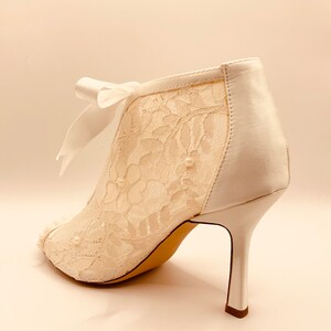 May include: White lace ankle boots with a satin back and a pearl embellishment. The boots have a delicate bow at the ankle and a high stiletto heel.
