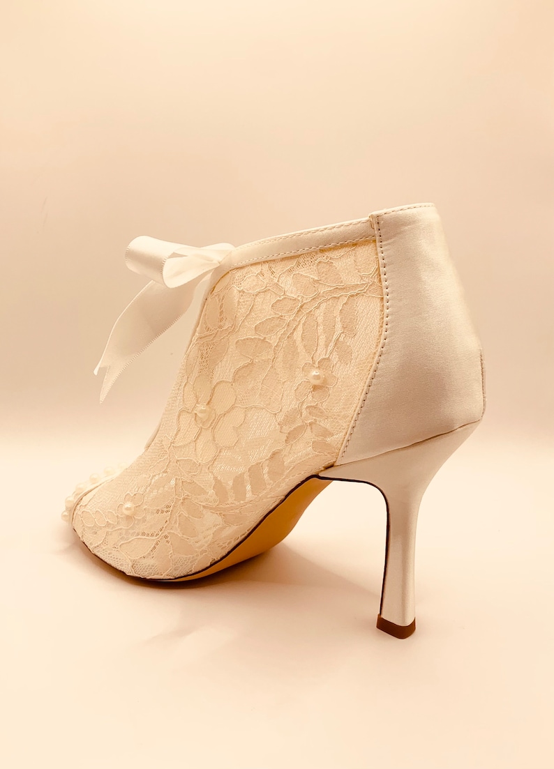 May include: White lace ankle boots with a satin back and a pearl embellishment. The boots have a delicate bow at the ankle and a high stiletto heel.