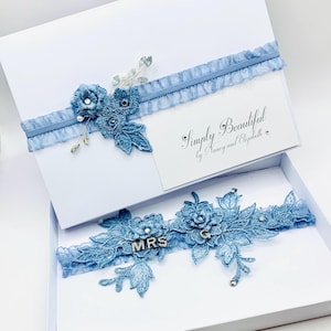 Op de afbeelding: Een blauwe kanten kousenband met een bloemenpatroon en het woord "MRS" in strass. De kousenband zit in een witte doos met een blauw lint en een kaartje met de tekst "Simply Beautiful by Nancy and Elizabeth".