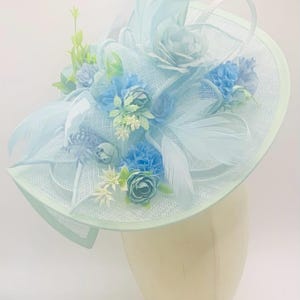 May include: A light blue hat with a wide brim and a large flower arrangement on top. The flowers are blue, white, and green. The hat is made of a delicate, mesh-like material.
