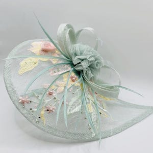 Peut inclure: Un chapeau bleu clair orné d'un motif floral. Le chapeau présente une base en résille avec des fleurs brodées roses et jaunes, des feuilles vert pâle, des perles et des cristaux. Un bouquet de tissu et de rubans orne le dessus.