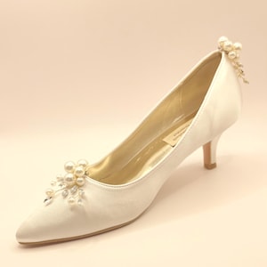 Puede incluir: Zapato de novia de satén marfil con punta y tacón bajo. El zapato está adornado con adornos de perlas y cristales en la puntera y el talón. Un diseño clásico y elegante para bodas y ocasiones especiales.