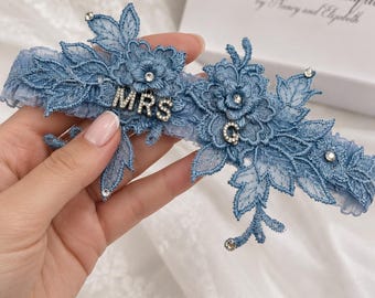 May include: A dusty blue lace garter featuring intricate floral embroidery, scattered clear rhinestones, and silver-toned metal lettering that spells out MRS and the letter G. The garter is crafted from gathered blue fabric with a delicate lace trim.