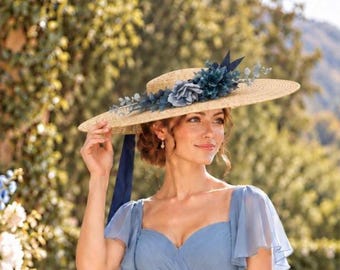 Puede incluir: Un sombrero de paja de ala ancha con copa plana, decorado con un arreglo de flores de tela azul oscuro y follaje verde. Una cinta larga de color azul oscuro cuelga del lateral del sombrero, que se lleva con un vestido azul claro.