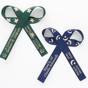 May include: Two fabric ribbons tied in loops, one dark green with copper star patterns and one navy blue with gold moon and star patterns. Each ribbon features printed text including names, dates, and the phrases In Loving Memory and Missed by All.