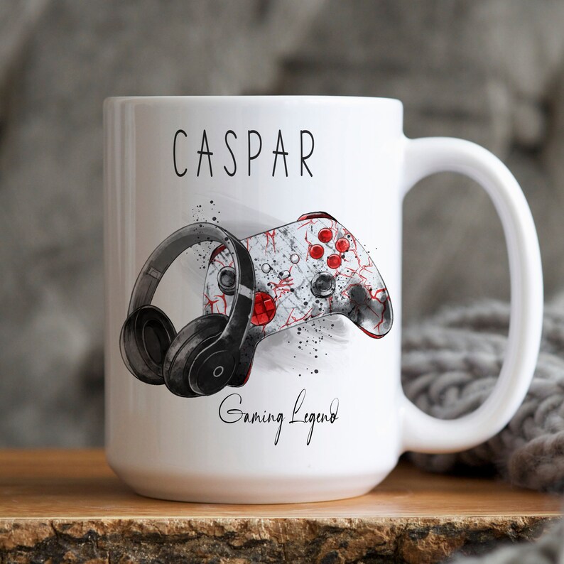 Pu&ograve; includere: Una tazza in ceramica bianca con un'illustrazione in bianco e nero di un controller da gioco e cuffie. La tazza ha il nome "CASPAR" e il testo "Gaming Legend" stampato.