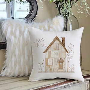 Peut inclure: Un coussin carré blanc avec une illustration à l'aquarelle d'une maison, d'un arbre et de fleurs. Le texte "NEW HOME" est imprimé en marron. Un coussin en fausse fourrure blanche et moelleuse est en arrière-plan.
