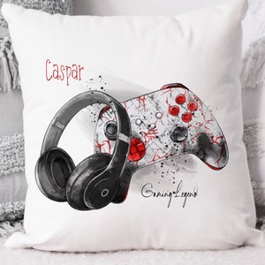 Pu&ograve; includere: Un cuscino bianco con un controller da gioco nero e rosso e cuffie nere. Il testo "Caspar Gaming Legend" &egrave; stampato sul cuscino.