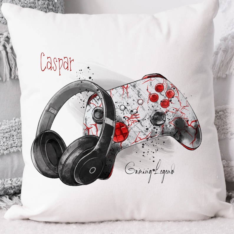 Pu&ograve; includere: Un cuscino bianco con un controller da gioco nero e rosso e cuffie nere. Il testo "Caspar Gaming Legend" &egrave; stampato sul cuscino.
