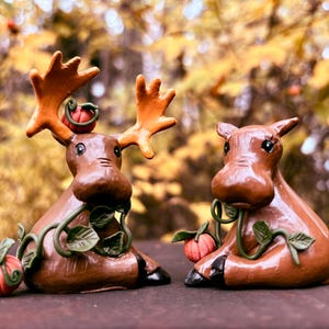 May include: Two brown moose figurines with orange antlers and small red pumpkins. One moose has large antlers, the other has small ears. Green vines with leaves and pumpkins decorate the moose figurines. The background is blurred.
