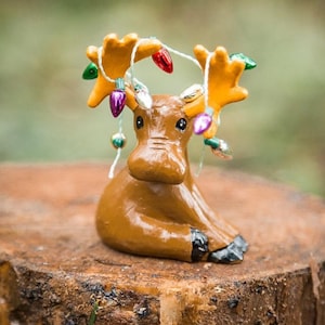 May include: A brown ceramic moose figurine with yellow antlers and colorful Christmas lights. The moose is sitting on a tree stump. The figurine is a festive holiday decoration.