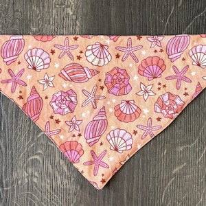 May include: A coral-colored pet bandana featuring a repeating pattern of seashells, starfish, and floral accents in shades of pink and white. The bandana is triangular in shape and made of fabric.