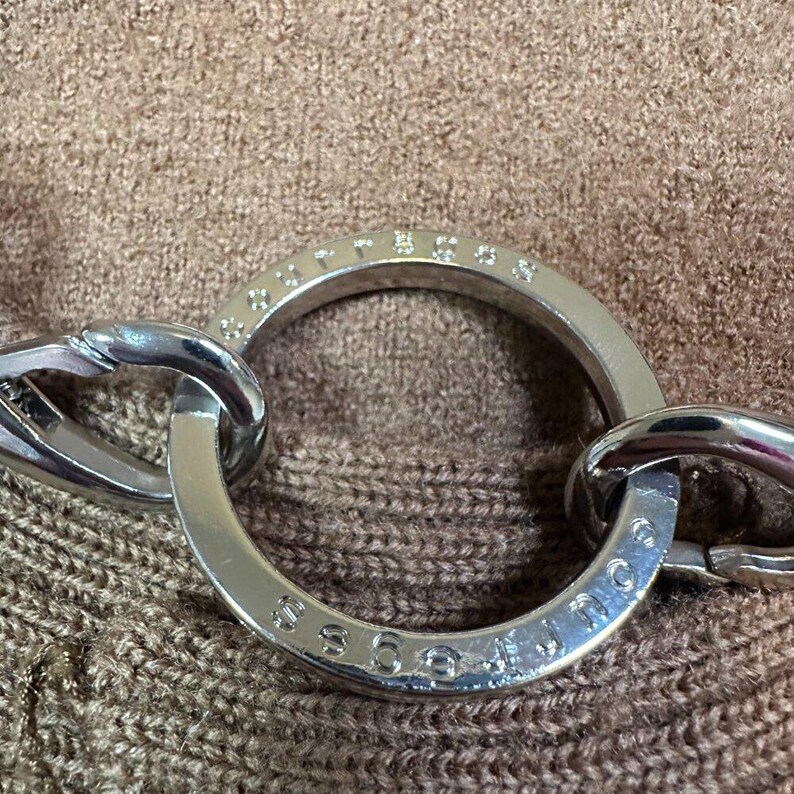 May include: Close-up of a silver-toned metal necklace with a large circular pendant. The pendant has embedded small crystals and the word "GUESS" engraved on it. The necklace is set against a brown textured background.