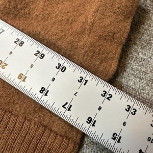 May include: Close-up of a brown fabric with a ruler laid across it. The ruler shows measurements in inches, with numbers from 15 to 33 visible. The fabric has a textured appearance.