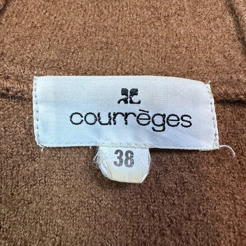 May include: Close-up of a brown fabric with a white label. The label features the brand name "courr&egrave;ges" in black font, along with a logo and the number "38". The fabric has a textured appearance.