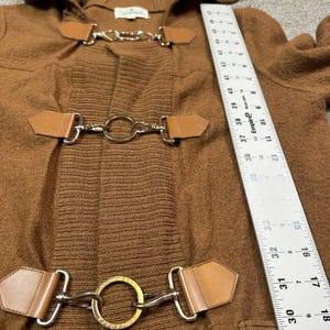 May include: A brown hooded coat with three sets of toggle closures. The closures feature silver metal hardware and brown leather accents. The coat has a ribbed knit section and a soft, textured fabric. A ruler is visible in the image.