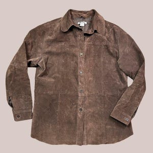 May include: A brown suede button-down shirt with long sleeves. The shirt has a collared neckline and is buttoned up the front. The suede material has a textured appearance. The shirt is a neutral color and suitable for various outfits.
