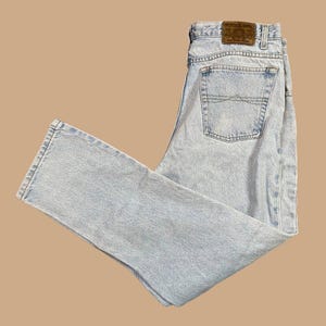 May include: Light blue denim jeans with a classic five-pocket design. The jeans feature a faded wash and a brown leather patch on the back. The jeans are folded and laid flat on a neutral background.