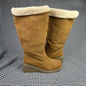 May include: A pair of tan suede boots with a cream-colored, fluffy trim at the top. The boots have a wedge heel and decorative stitching. The boots are approximately 12 inches tall.