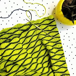 Puede incluir: Una manta de crochet verde y negra con un patr&oacute;n ondulado. La manta se est&aacute; trabajando con un gancho de crochet.