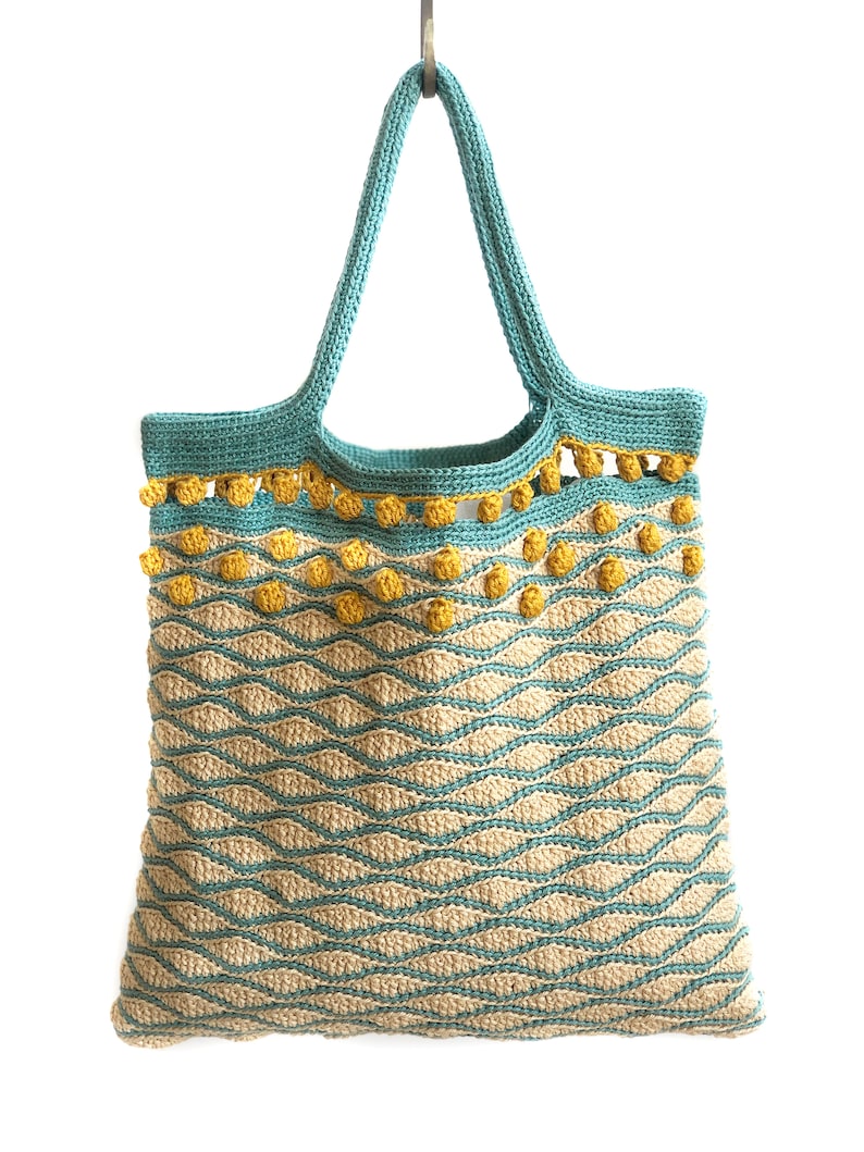 Puede incluir: Una bolsa de mano de crochet de color azul verdoso y beige con pompones amarillos alrededor del borde superior. La bolsa tiene un patr&oacute;n ondulado.