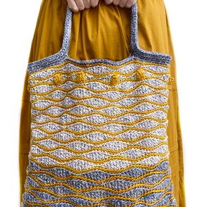 Puede incluir: Un bolso de mano de crochet con un patr&oacute;n ondulado en tonos azules y amarillos. El bolso tiene una correa gruesa de crochet y pompones amarillos.
