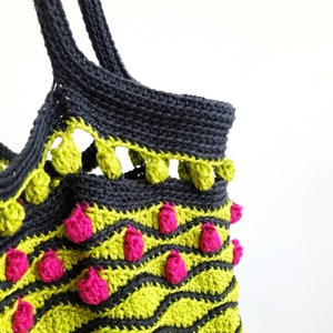 Puede incluir: Una bolsa de mano de crochet negra con ribete de pompones verdes y rosas. La bolsa tiene un patr&oacute;n ondulado y est&aacute; hecha con hilo grueso.
