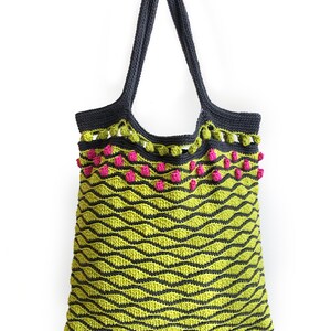 Puede incluir: Una bolsa de mano de crochet verde y negra con pompones rosas alrededor de la parte superior.