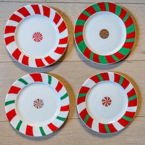 May include: Four white ceramic plates with red and green candy cane stripes. Each plate has a small, stylized candy design in the center.