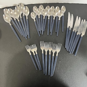 Puede incluir: Un juego de cubiertos de acero inoxidable con mangos azul marino. El conjunto incluye cucharas, tenedores y cuchillos. La cubertería está dispuesta sobre una superficie oscura, mostrando el diseño elegante y el contraste de color.