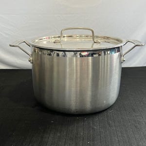 May include: A stainless steel stockpot with a matching lid. The pot has two side handles and a loop handle on the lid. The pot is sitting on a black surface.
