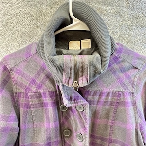 May include: A gray and purple plaid jacket with a gray ribbed collar and a zipper closure. The jacket has a button-up front and is hanging on a white hanger.